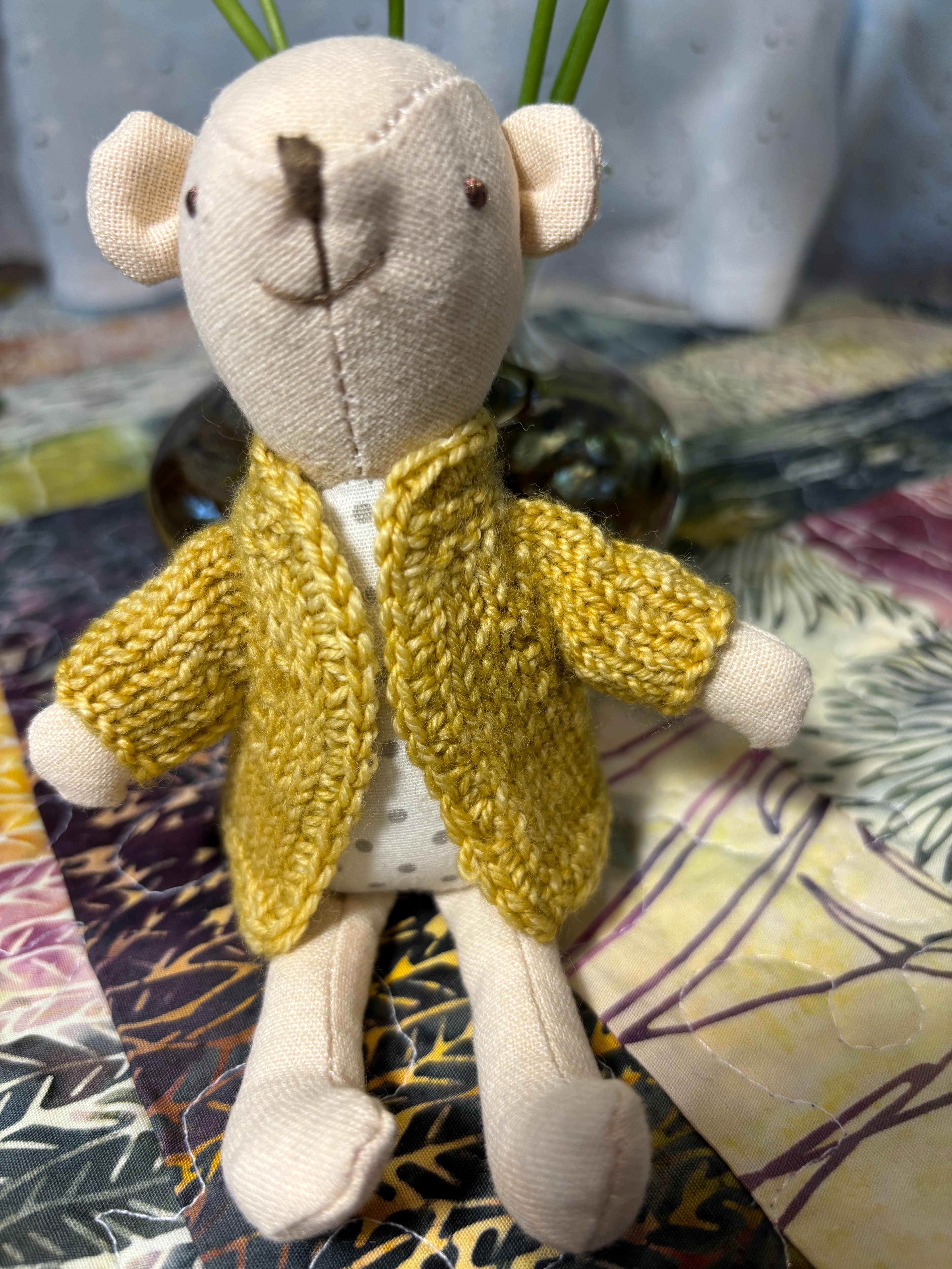 A small cloth doll wearing a mustard hand-knit cardigan, styled on a quilt.