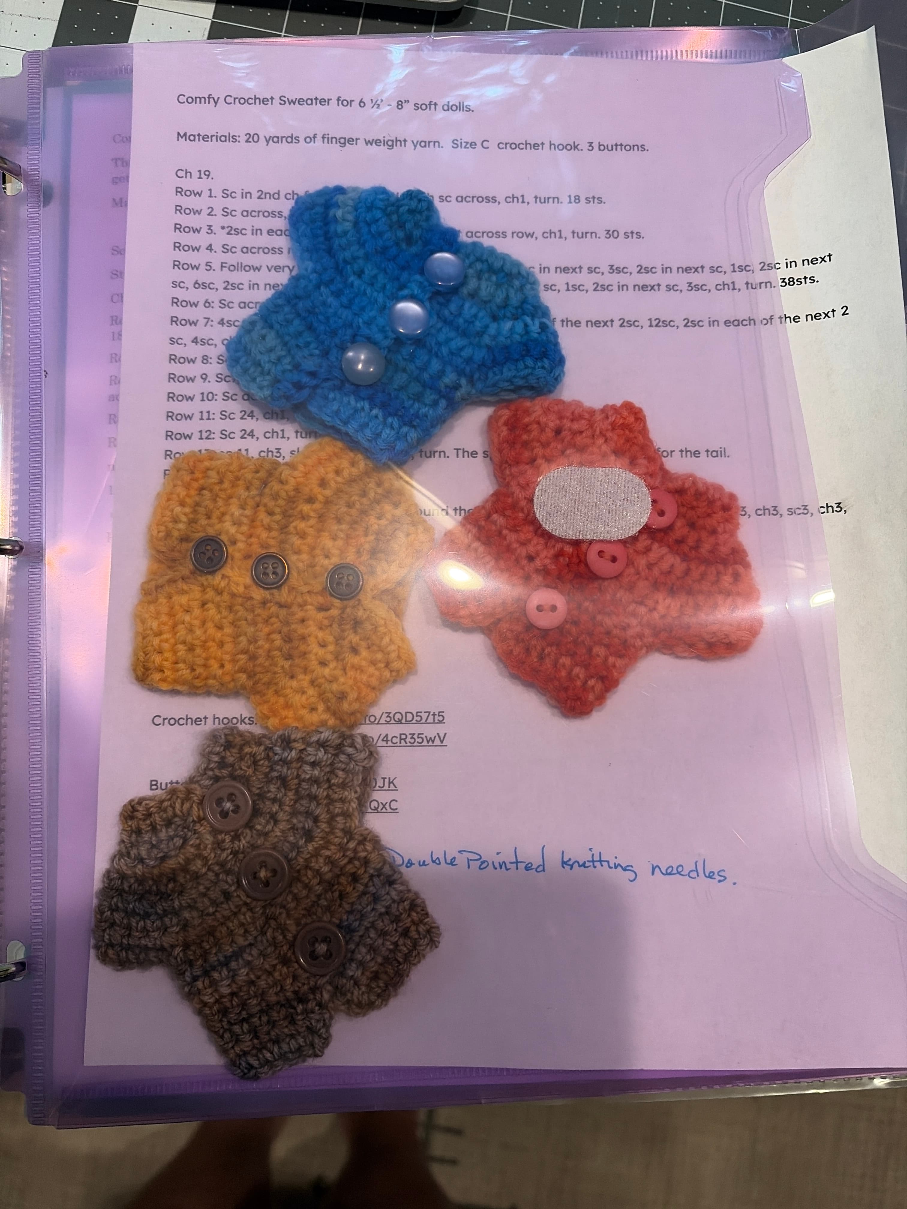 Four small crochet doll sweaters in coral, blue, gold and brown with buttons, resting on the crochet pattern sheet.