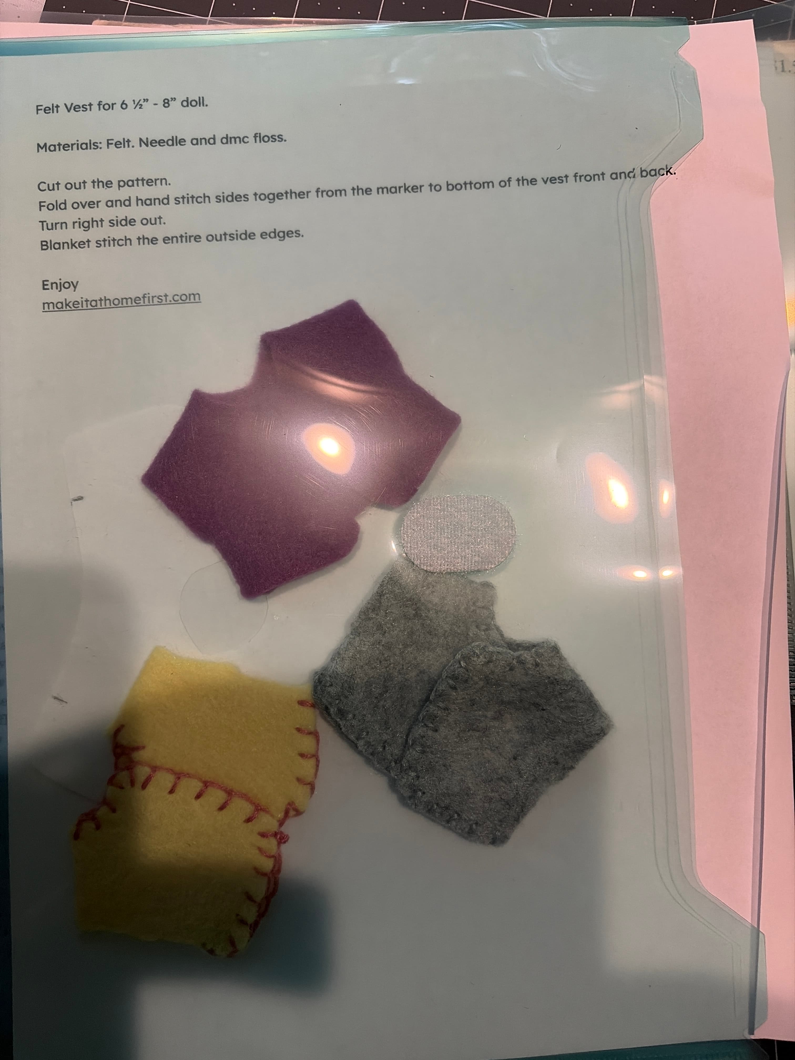 Three small felt doll vests in grey, plum and yellow with hand-stitched edges, on the felt vest pattern.