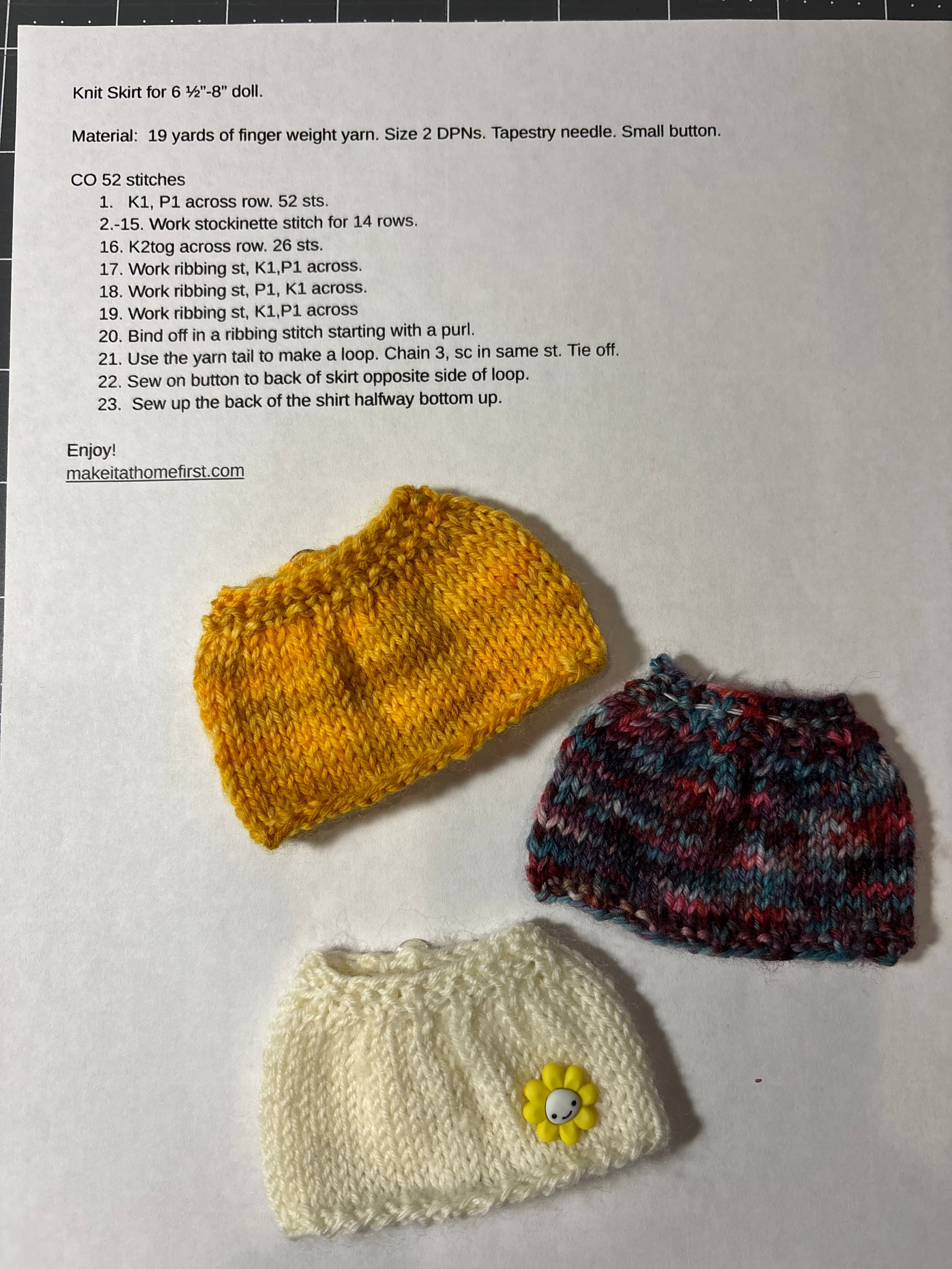 Three small knit doll skirts in berry, gold and cream, one with a yellow flower button, on the knit skirt pattern.